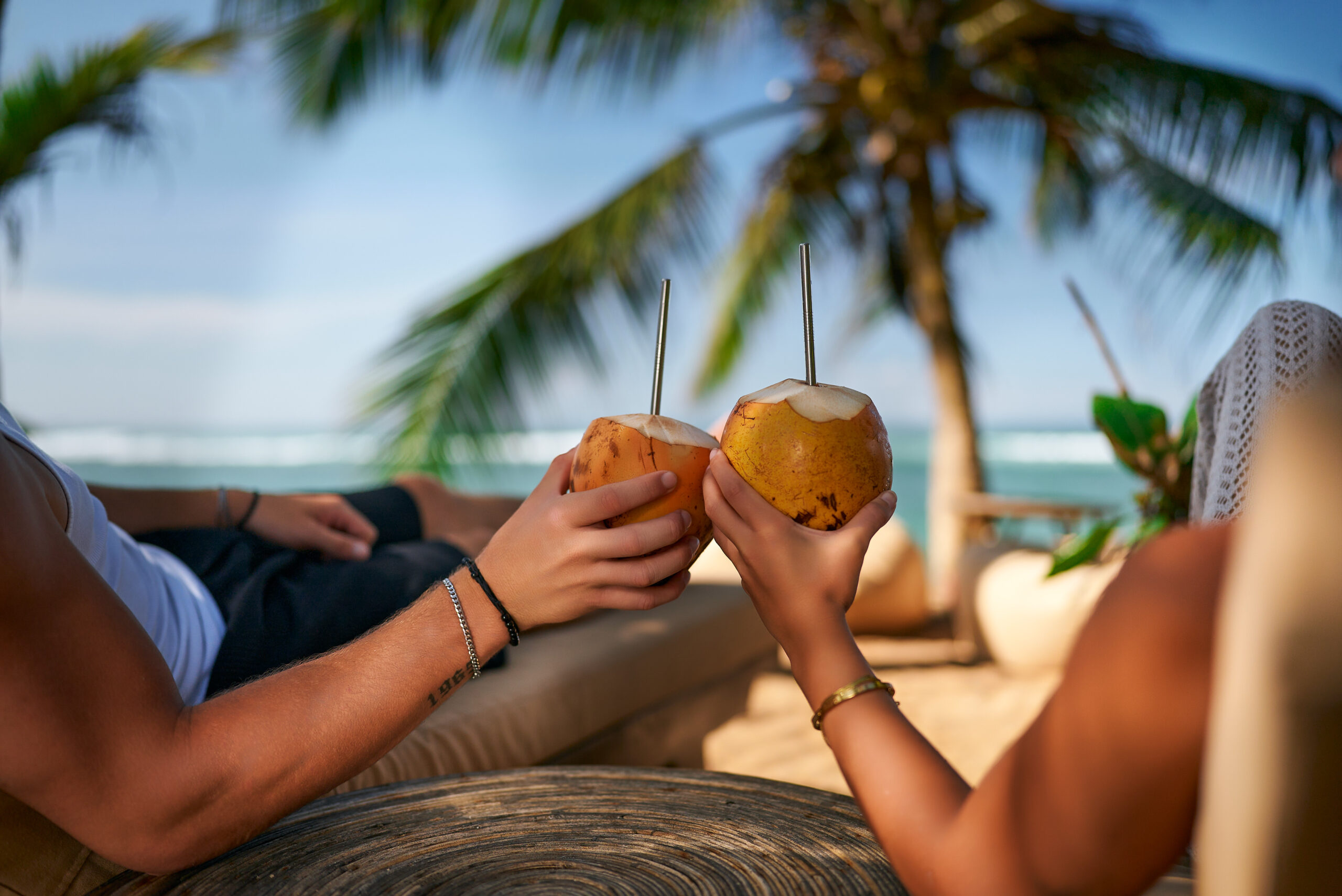 Romantic tropical getaway, pair sips natural drinks under palm tree. Couple relaxing on beach lounge chairs, enjoying fresh coconut water. Ocean view, sunny day, luxury resort, leisure lifestyle.